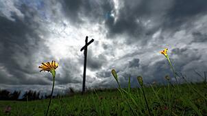 Blumen stehen auf einer Wiese vor einem Wegkreuz unter dichten Regenwolken. (Symbolbild) Eine 51-Jährige Frau stürzt beim Wandern im ...