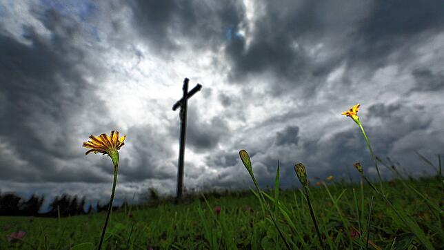 Blumen stehen auf einer Wiese vor einem Wegkreuz unter dichten Regenwolken. (Symbolbild) Eine 51-Jährige Frau stürzt beim Wandern im ...