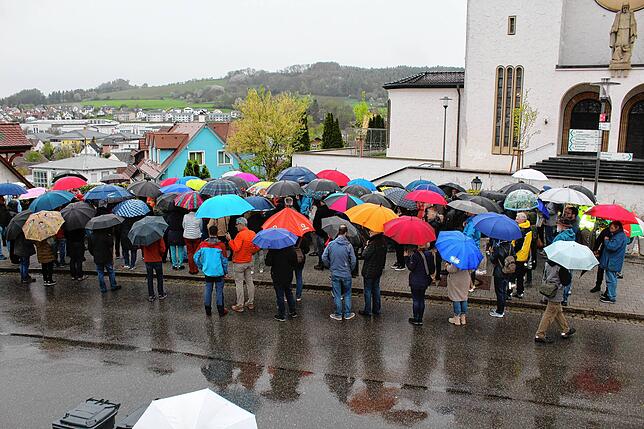 Viele beschirmte Gäste nehmen bei Regenwetter am Informations-Spaziergang teil. Hier stehen sie an der Mauer zwischen Hauptstraße und ...