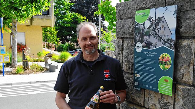 Andreas Bohnenstengel, Chef der Süßmostkelterei Wilhelm Auer, mit Apfelwein am Schild des Heimatpfads der Gemeinde, bei dem die Kelterei ...