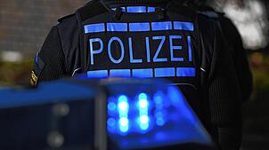Das Logo der Polizei auf der Dienstkleidung eines Polizisten der vor einem Blaulicht steht. (Symbolbild)