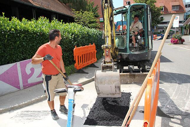 Mark Laski (links) verteilt mit einem Besen Asphalt, das Richard Walny mittels Bagger auf die zu füllende Stelle ablädt.