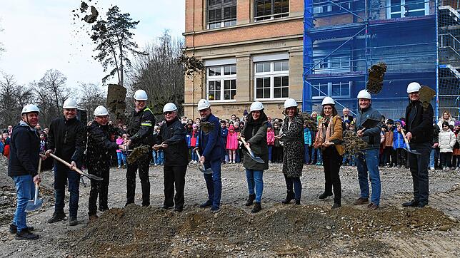 Baubeteiligte und Bürgermeisterin machen den Spatenstich für das Zwischengebäude der Grundschule. Alle Schüler schauen zu.