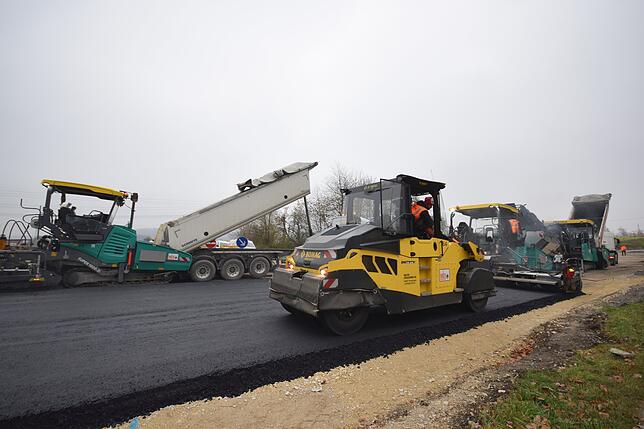 Hinten beliefert ein Laster den Beschicker, vorne fährt eine Walze und verdichtet aufgebrachten Asphalt.