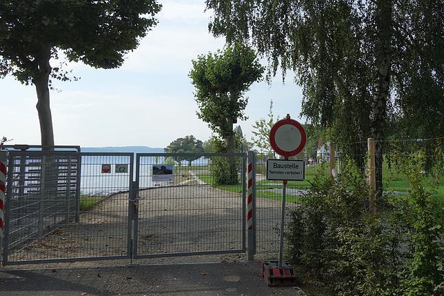 Rund um den Uferpark zieht sich ein Maschendrahtzaun mit Holzpfählen. Am östlichen Ende ist schon das Drehkreuz eines Ausgangs installiert.