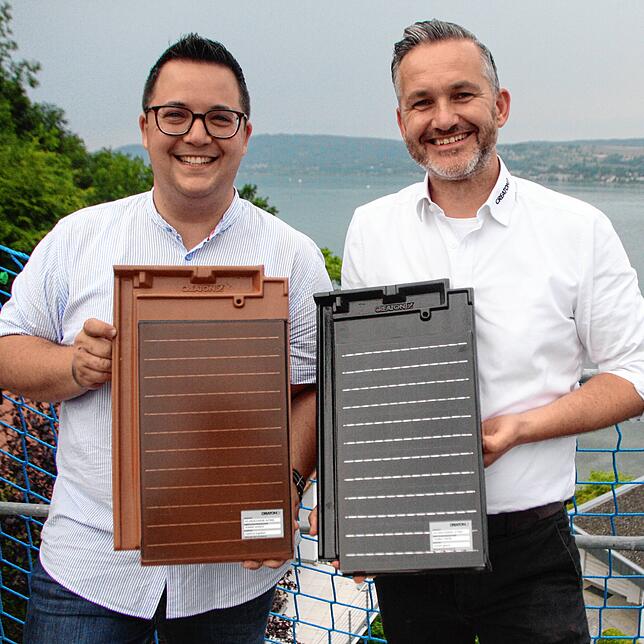 Dario Sauchelli und Robert Kirchner präsentieren die Solarziegel. Sauchelli hält das Modell in Rot in der Hand, Kirchner das in Dunkel.