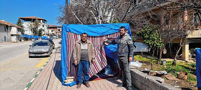 Zwei Männer stehen in Antakya vor ihren wenigen Habseligkeiten, die sie mit Decken und Planen schützen. Seit dem Erdbeben leben viele ...