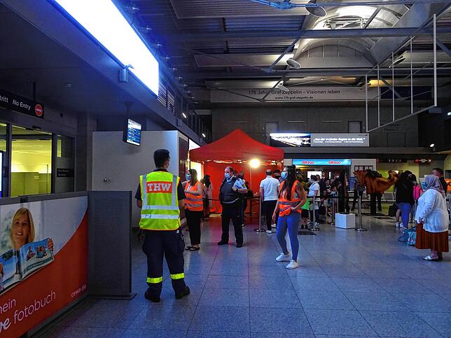 Mitarbeiter des Landratsamts, hier mit roter Warnweste, und des Technischen Hilfswerks sprechen Reisende im Ankunftsbereich an und ...