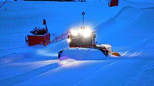 Mit dem mächtigen Schild schiebt die mit einer Seilwinde ausgestattete Pistenraupe in Fahl Schnee vom Rand auf die Skipiste. Ein ...