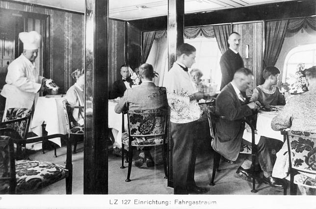 Wie in einem erstklassigen Hotel: Der Fahrgastraum von LZ 127. Links ein Koch mit Mütze, in der Mitte bedient ein Kellner in Weiß die Gäste.