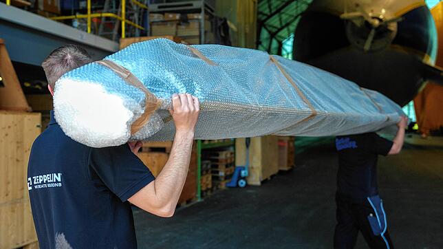 Eine Lieferung Längsträger: Das Bauteil, das die beiden Mitarbeiter hier geschultert haben, wird im Zeppelinhangar sicher deponiert und ...