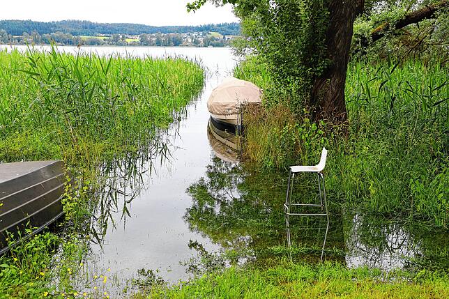 Die Beine eines Stuhls, der auf einer gefluteten Wiese steht, sind teilweise vom Wasser bedeckt. Kalte Füße aufgrund des Hochwasser ...