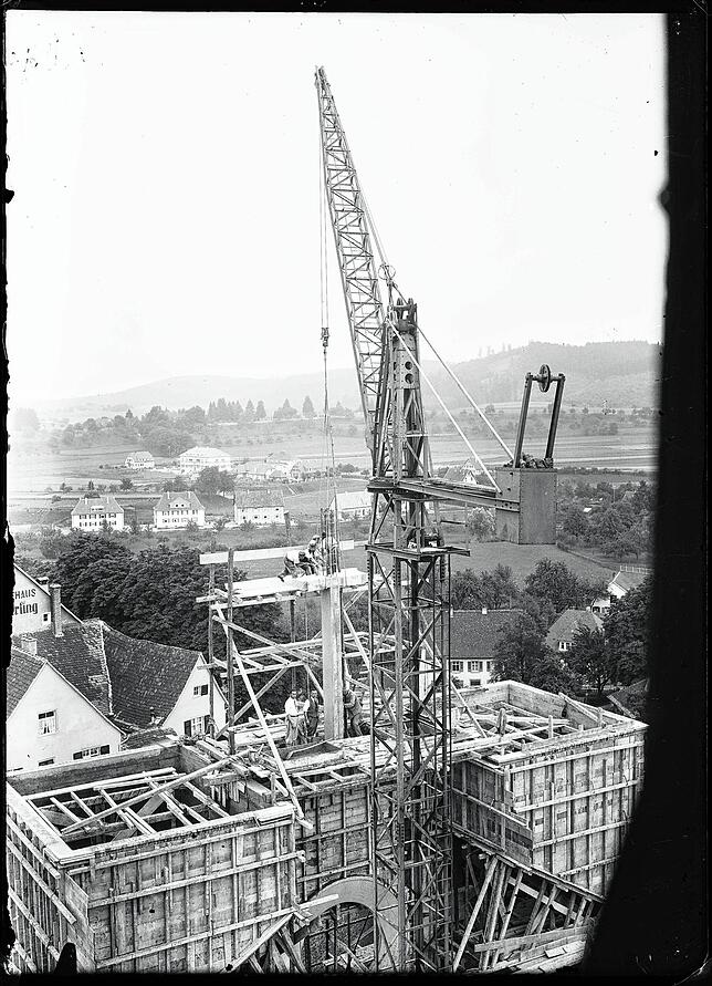 Auf dem Gerüst beim Kran stehen Arbeiter, um mithilfe des Krans ein Stützsegment in die Wand einzulassen. Am unteren Bildrand ist das ...
