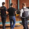 Der Angeklagte wird von zwei Justizbeamten in den Verhandlungsraum geführt. An seinen Händen und Füßen trägt er Fußfesseln.