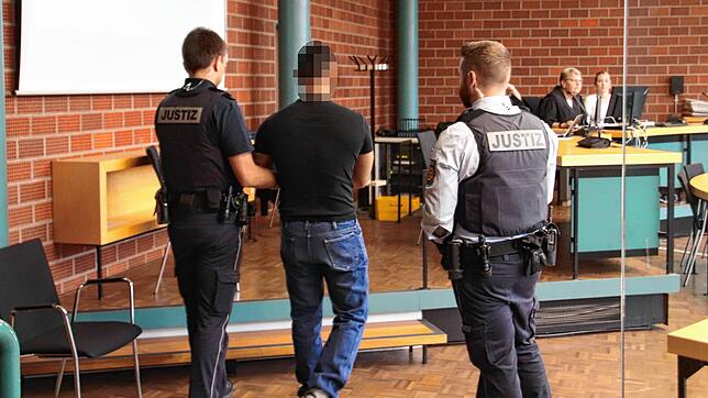 Der Angeklagte wird von zwei Justizbeamten in den Verhandlungsraum geführt. An seinen Händen und Füßen trägt er Fußfesseln.