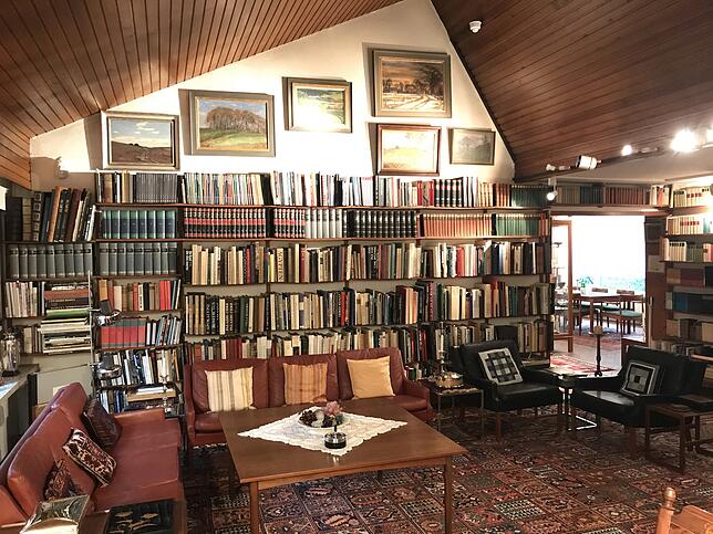 Das Wohnzimmer mit Couch-Ecke, Tisch und der zentralen Bücherwand. Darüber Landschaftsgemälde. Rechts hinten der Durchgang zum Esszimmer ...