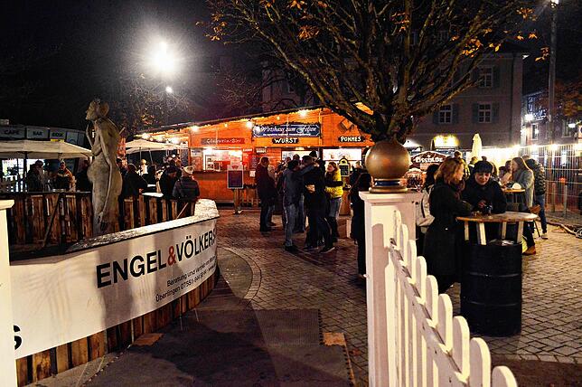 Besucher stehen in kleinen Gruppen an den Stehtischen. Sie trinken Glühwein oder Punsch, essen Pommes oder eine Wurst – und freuen ...