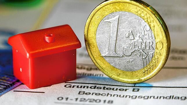 Eine Euro-Geldmünze sowie ein Spielzeughaus stehen auf einem Abgabenbescheid für die Entrichtung der Grundsteuer.