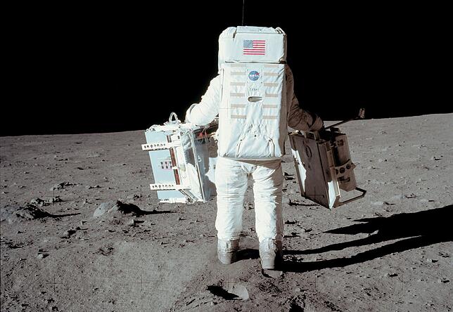 Edwin Aldrin auf dem Mond von hinten: Er schleppt zwei Messgeräte. Gut sichtbar ist sein Überlebensrucksack, der Sauerstoff zum Atmen ...