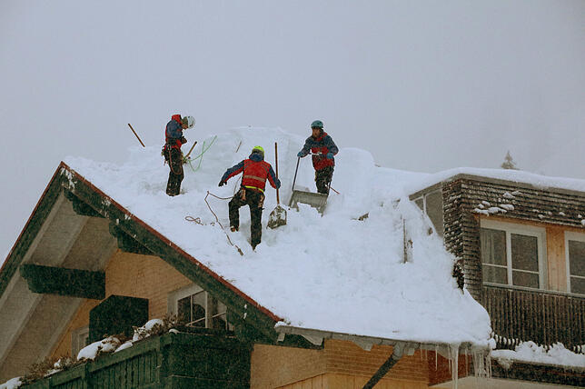 Einsatzkräfte stehen in der tief verschneiten Ortschaft auf den Hausdächern und schaufeln Schnee herunter.