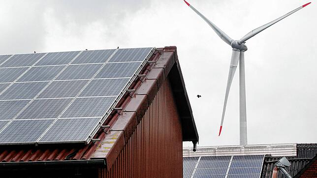 Hinter den Dächern eines Bauernhofes mit Photovoltaik produziert eine Windkraftanlage Öko-Strom. Diese Art Strom zu erzeugen ist nicht ...