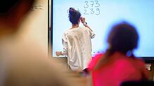 Eine Lehrerin schreibt eine Aufgabe auf eine digitale Schultafel im Klassenraum einer vierten Klasse einer Grundschule. Gerade in ...
