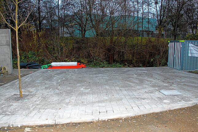 Noch ein leerer Platz mit Betonpflastersteinen. Dort wird eine Tischtennisplatte stehen. Hinter den Bäumen eine Sporthalle des ...