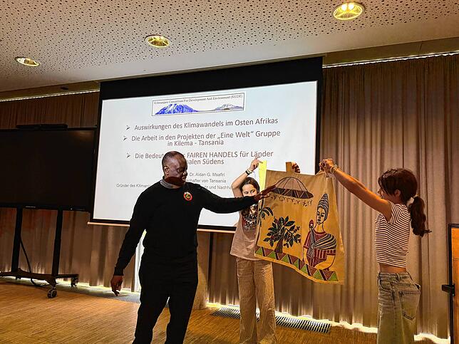 Pater Aidan überreicht ein Kunstwerk aus Stoff an die Robert-Schumann-Realschule.
