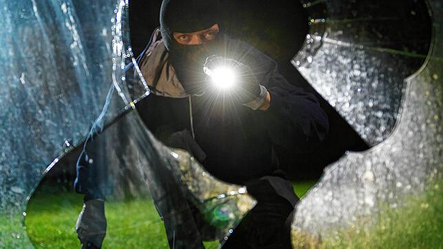 Symbolbild: Ein vermumter Mann mit Taschenlampe blickt in einer gestellten Szene durch eine eingeschlagene Fensterscheibe.