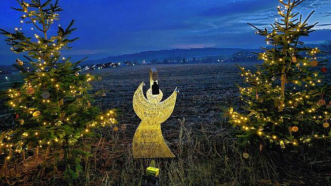 Ein Engelsschild mit Text und Melodie zu „Stille Nacht“, flankiert von zwei Weihnachtsbäumen.