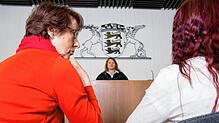 Eine Prozessbegleiterin sitzt in einem Gerichtssaal neben einer Zeugin. Sie helfen den Opfern, auch im Angesicht ihres Peinigers ruhig ...