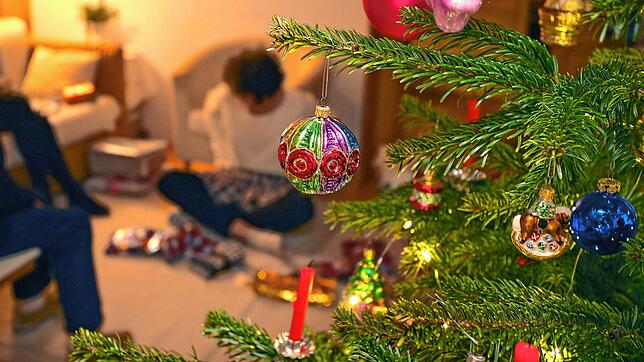 Eine Familie packt hinter einem geschmücktem Christbaum Weihnachtsgeschenke aus. Dazu darf es auch dieses Jahr wieder kommen.