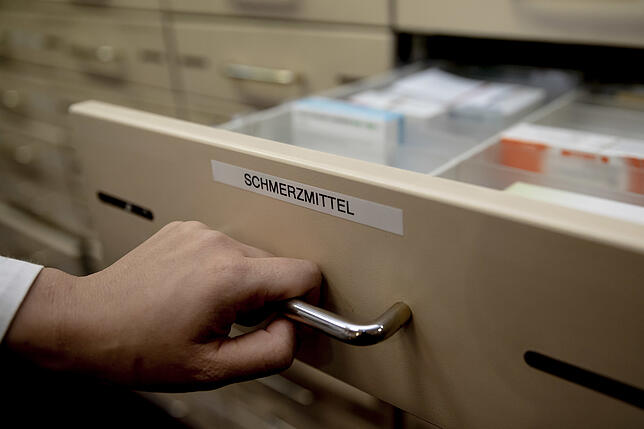 Eine Apothekerin öffnet in einer Apotheke eine mit “ Schmerzmittel“ beschriftete Schublade.