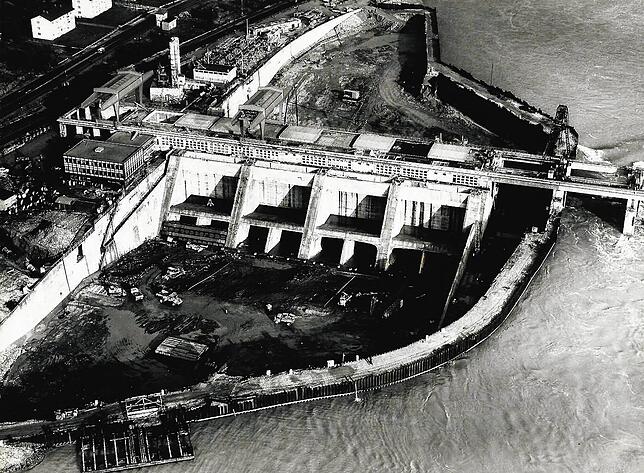 Tief reicht die Baugrube beim Bau des Rheinkraftwerkes bis auf den Rheingrund. Viele Meter über der Baustelle strömen die Fluten des Rheins.