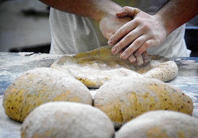 Ein Bäcker formt am Morgen in der Backstube auf einer mit Mehl bestäubten Arbeitsplatte den Teig für Kürbiskernbrötchen. Brot und ...