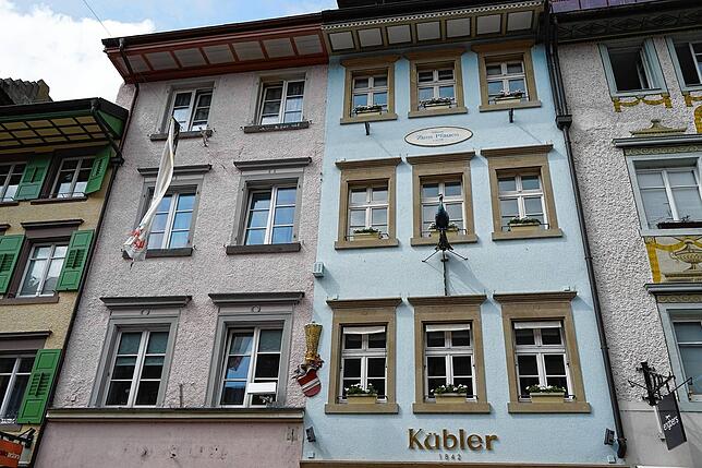 Links rosa, rechts hellblau – aber einst waren diese beiden Gebäude ein großes Haus. Das Wappen in der Mitte der Fassade zeigt es.