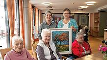 Annette Loll vom Kulturring und Martina Meier (stehend von links) mit einem Werk von Ingrid Veit. Die Bewohnerinnen freuen sich ...