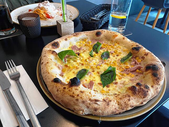 Auf der anderen Tischseite gibt es eine Pizza Mimosa mit Sahne, Schinken und Mais.