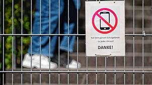 Ein Schild mit einem durchgestrichenen Smartphone und der Aufschrift „Auf dem ganzen Schulgelände – zu jeder Uhrzeit! DANKE!“ hängt in ...