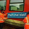 BOB-Mitarbeiter kleben einen blauen Sticker auf die rote Bahn. Darauf zu lesen: &bdquo;Mach mal blau!&ldquo;