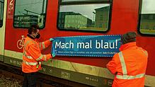 BOB-Mitarbeiter kleben einen blauen Sticker auf die rote Bahn. Darauf zu lesen: &bdquo;Mach mal blau!&ldquo;