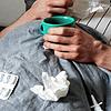 Ein Mann liegt mit Taschentüchern, Teetasse, Nasenspray und Tabletten im Bett (gestellte Szene).