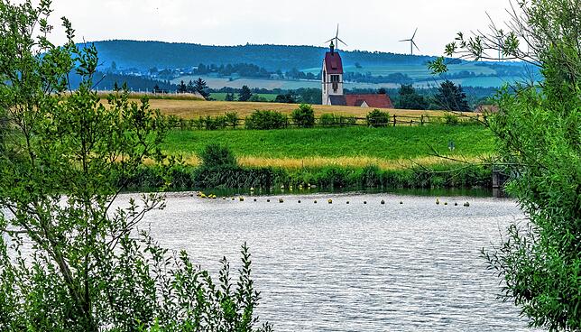 Der See ist von Feldern und Wiesen umgeben. Im Hintergrund ist der Sunthauser Kirchturm zu sehen.
