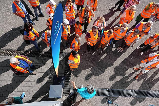 Besucher versammeln sich beim Tag der offenen Tür des Rheinkraftwerks Rheinfelden in orangenen Warnwesten vor einem Banner mit dem neuen ...