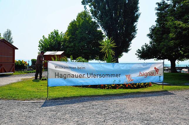 „Willkommen beim Hagnauer Ufersommer“ steht auf dem Banner. Im Hintergrund stehen Hütten. Außerdem wässert ein Mitarbeiter die Pflanzen ...