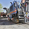 Eine riesige Fräsmaschine mit Kettenantrieb trägt die oberste Schicht Asphalt an den zuvor markierten Schadstellen ab. Das Material wird ...