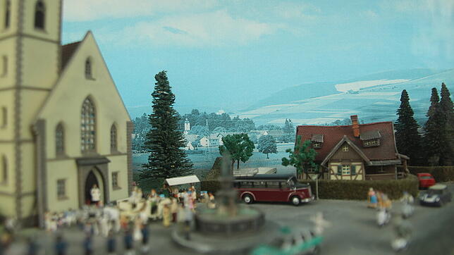 Ein bemalter, durch die Sonne mittlerweile etwas verblasster, Hintergrund. Im Vordergrund die Kirche und Brunnen.