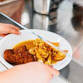Eine Grundschülerin in Freiburg nimmt einen Teller mit Schnitzel, Kartoffeln und Sauce vom Tresen der Essensausgabe einer Schulmensa. Ab ...