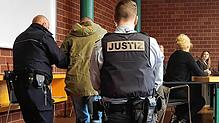 Der Angeklagte, der Hand- und Fußfesseln trägt, wird von Justizbeamten zu seinem Platz im Gerichtssaal geführt, wo eine Dolmetscherin ...