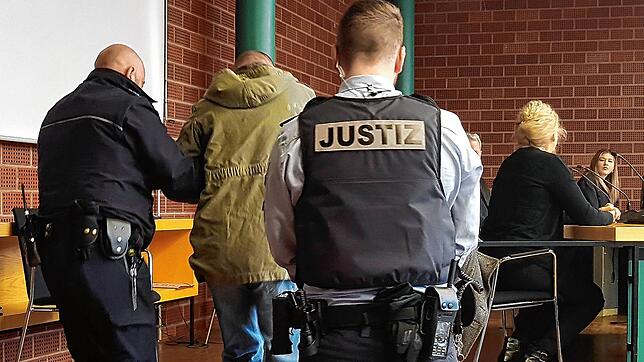 Der Angeklagte, der Hand- und Fußfesseln trägt, wird von Justizbeamten zu seinem Platz im Gerichtssaal geführt, wo eine Dolmetscherin ...